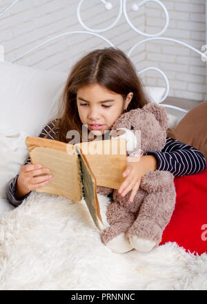 Kleines Mädchen auf dem Bett liegend mit ihren Teddybären und lesen Sie ein Buch vor dem Schlafengehen Stockfoto