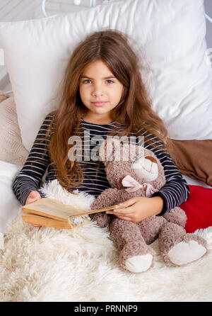 Kleines Mädchen auf dem Bett liegend mit ihren Teddybären und lesen Sie ein Buch vor dem Schlafengehen Stockfoto