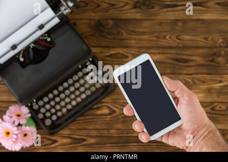 Nicht identifizierbarer Mann mit weissen smart phone neben Black Metal vintage Schreibmaschine und Kleine rosa Blumen Stockfoto