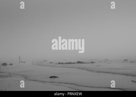 Einzelne Person an einem Strand mit Wellen auf Sand und Felsen im Hintergrund in Schwarzweiß Stockfoto