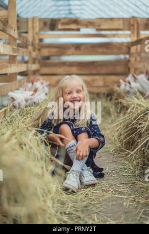 Glückliches Kind sitzt auf dem Boden in der Nähe von Ziegen auf der Farm und Kamera Stockfoto