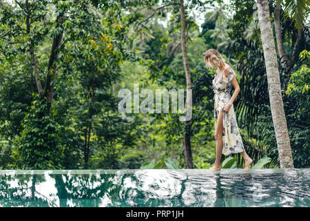 Attraktive blonde Frau im Kleid zu Fuß in der Nähe von Schwimmbad mit grünen Pflanzen auf Hintergrund, Ubud, Bali, Indonesien Stockfoto