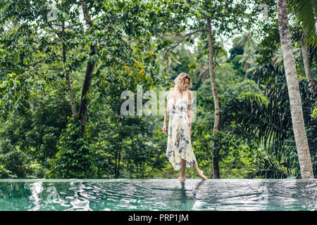 Attraktive blonde Frau im Kleid zu Fuß in der Nähe von Schwimmbad mit grünen Pflanzen auf Hintergrund, Ubud, Bali, Indonesien Stockfoto