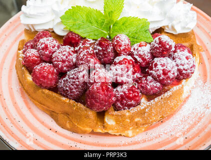 Leckere belgische Waffeln mit frischen Himbeeren und Schlagsahne mit Mandel- Flocken. Mit Minze und Puderzucker eingerichtet, auf orange Platte. Del Stockfoto