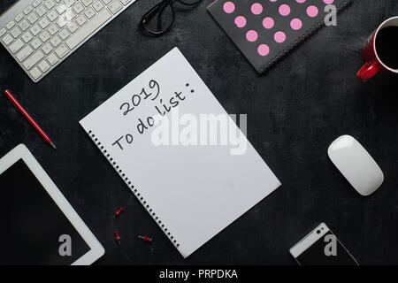 Notiz Buch, Smart Phone und andere Verbrauchsmaterialien mit Tasse Kaffee mit Text 2019, um Liste zu tun. Holz- schwarz Büro Schreibtisch Tisch mit Tischplatte Blickwinkel. Stockfoto