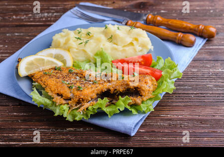 Gebratenen Fischen auf grünem Salat mit Kartoffelpüree, Tomate und Zitronenscheiben in blauen Platte auf dunklen Holzmöbeln im Landhausstil Hintergrund. Köstliches Abendessen. Stockfoto