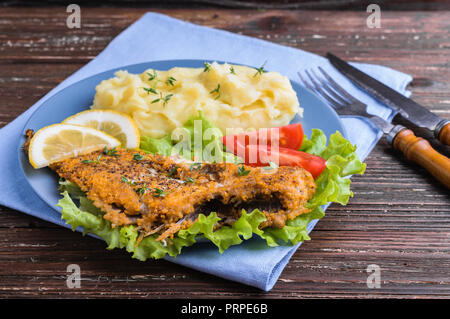 Gebratenen Fischen auf grünem Salat mit Kartoffelpüree, Tomate und Zitronenscheiben in blauen Platte auf dunklen Holzmöbeln im Landhausstil Hintergrund. Köstliches Abendessen. Stockfoto