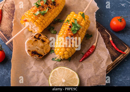 Gegrillte Maiskolben. Köstliche Sommer Snack. Mit Petersilie, Chili peppers, Kalk und gegrillter Knoblauch. Auf alten hölzernen Schneidebrett mit Pergament pap serviert. Stockfoto