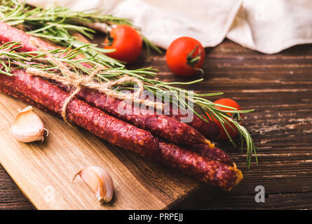 Kabanosy - Geräucherte traditionelle polnische Würstchen auf Holz Schneidebrett. Mit Rosmarin, Kraut, Knoblauchzehen und Tomaten. Auf rustikalen Holztisch Stockfoto