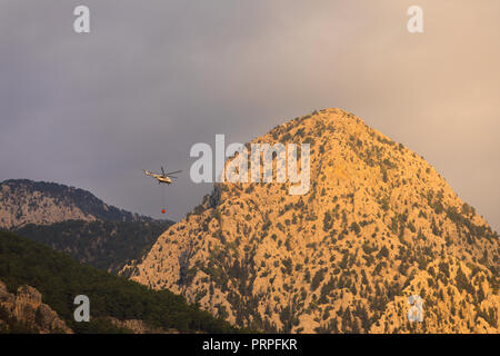 Ein Brand Hubschrauber mit einem vollen Korb von Wasser fliegt ein waldbrand vor dem Hintergrund der Berge zu löschen, in hellen orange sun Farbe lackiert Stockfoto