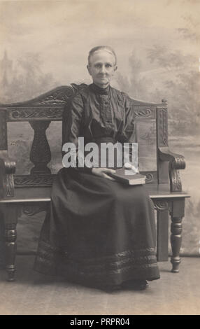 * Vintage makabren Foto eines Toten ältere Frau setzte sich auf eine Bank, die ein Buch. Nähen sichtbar für den Mund und die Leichenstarre sichtbar auf Ihre Hände. Stockfoto