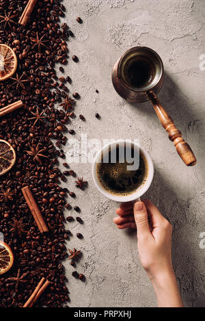 7/8 Schuß von Frau mit Tasse Kaffee auf die Betonoberfläche mit verschüttetem Kaffee Bohnen und Gewürze Stockfoto