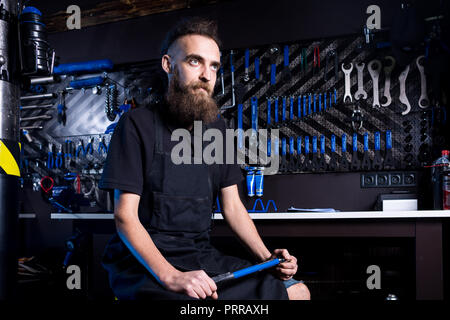 Portrait von Small Business Owner junger Mann mit Bart. Guy Fahrradmechaniker workshop Arbeiter sitzen mit Werkzeug in der Hand in einem schwarzen Kleide Stockfoto