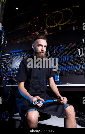 Portrait von Small Business Owner junger Mann mit Bart. Guy Fahrradmechaniker workshop Arbeiter sitzen mit Werkzeug in der Hand in einem schwarzen Kleide Stockfoto
