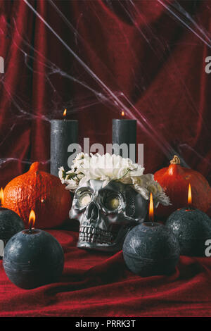 Silber Halloween Totenkopf mit Blumen, schwarze Kerzen und Kürbisse auf rotem Tuch mit Spinnennetz Stockfoto