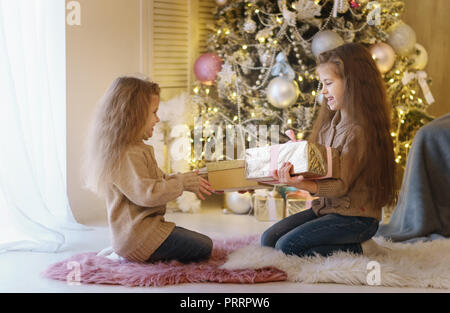 Zwei kleine Schwestern warten auf Weihnachten. Die Mädchen sitzen mit Weihnachtsbaum, lächelnd und Austausch von präsentiert. Stockfoto