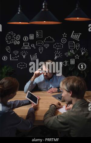 Geschäftsfrau etwas an der Tafel zu Ihrem Partner und Sitzen am Tisch, mit Hand gezeichnete Geschäft Symbole über den Köpfen Stockfoto