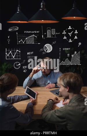 Geschäftsfrau etwas an der Tafel zu Ihrem Partner und Sitzen am Tisch, mit Business Icons über den Köpfen Stockfoto