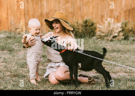 Glückliche Mutter in Hut und kleinen Sohn petting inländischen Ziege auf grünem Gras an der Landschaft Stockfoto