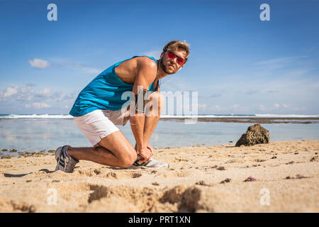 Sportler mit Smartphone armband in Sonnenbrille auf sneakers Schnürsenkel binden am Strand Stockfoto