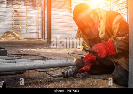 Ein Erbauer Schweißer in Braun Arbeitskleidung Schweißnähte ein Metall Produkt mit arc welding Maschine in der Fabrik, eine lose Werkzeuge und constraction, helle lig Stockfoto