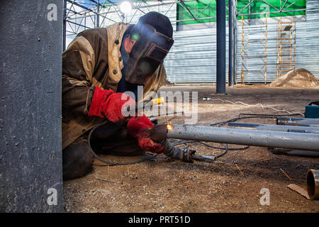 Ein Erbauer Schweißer in Braun Arbeitskleidung Schweißnähte ein Metall Produkt mit arc welding Maschine in der Fabrik, eine lose Werkzeuge und constraction, helle lig Stockfoto