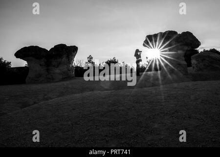 Nicht erkennbare Fotografin Silhouette bei Sonnenuntergang mit Sun Ray Diffraction gegen die Sonne in der Nähe des Steinernen Pilze natürliches Phänomen im Süden Bulgariens, in der Nähe der Stadt Kardschali, Schwarz und Weiß Stockfoto