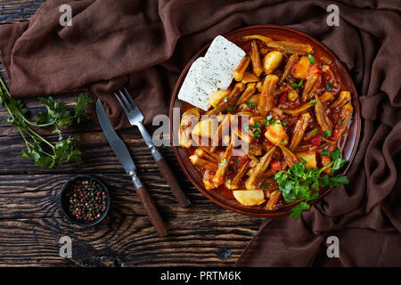 Leckere griechische okra Eintopf mit Kartoffeln mit Feta Käse auf einem Steingut Teller serviert auf einem rustikalen Tisch. Gabel, Messer, Petersilie und braunen Tuch an der Stockfoto