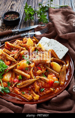 Griechische okra Tomate Eintopf mit Kartoffeln mit Feta Käse auf einem Steingut Teller serviert auf einem rustikalen Tisch, griechisch Rezept. Gabel, Messer, Petersilie am Stockfoto