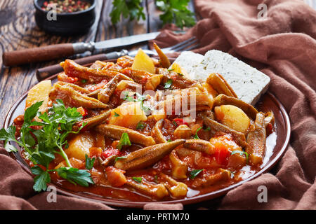 Geschmorte okra mit Tomaten und Kartoffel, serviert mit Feta Käse auf einem Steingut Teller auf einem rustikalen Tisch, griechisch Rezept. Gabel, Messer, Petersilie am Stockfoto