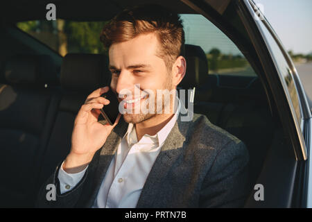 Portrait von erfolgreichen Mann 30 s im klassischen Anzug Reden auf dem Smartphone während zurück sitzen in der Business Class Auto Stockfoto