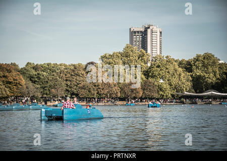 Zwei Männer in einem Tretboot auf dem Serpentine im Londoner Hyde Park und der Park Lane Hilton im Hintergrund Stockfoto