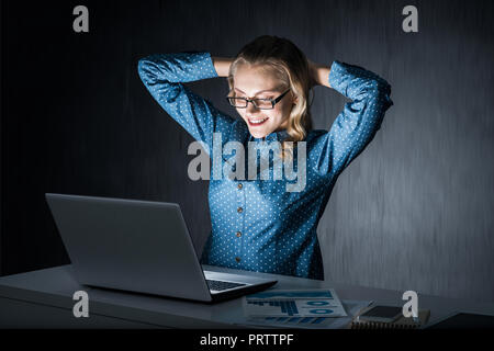 Attraktive Blondine Arbeiten am Laptop im dunklen Büro. Mixed Media Stockfoto