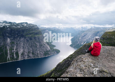 Majestic Trolltunga - eine von Norwegens spektakulärer Anblick. Mädchen in der roten Jacke sitzt auf Felsen und blickt auf Ringedalsvatnet See Stockfoto