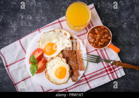 Englisches Frühstück - Speck, Rührei, Bohnen und Orangensaft. Auf weiße Platte mit in Scheiben geschnittenen Tomaten und Basilikum serviert. Dark grunge konkrete Tabelle b Stockfoto