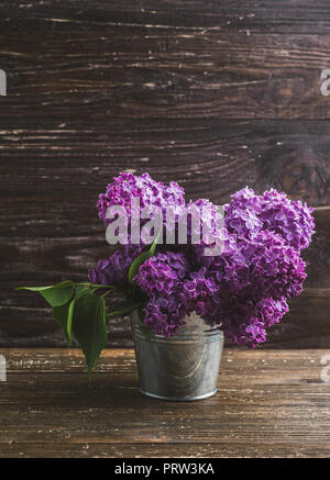 Blumenstrauß aus Lila lila Blüten in kleinen dekorativen Dose Eimer. Dunkel Braun Holz- Hintergrund. Feder romantische Blumen Dekoration. Stockfoto