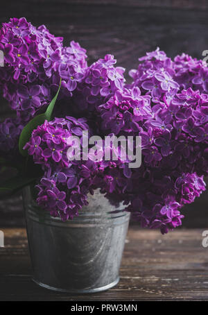 Blumenstrauß aus Lila lila Blüten in kleinen dekorativen Dose Eimer. Dunkel Braun Holz- Hintergrund. Feder romantische Blumen Dekoration. Close Up. Vintage fi Stockfoto