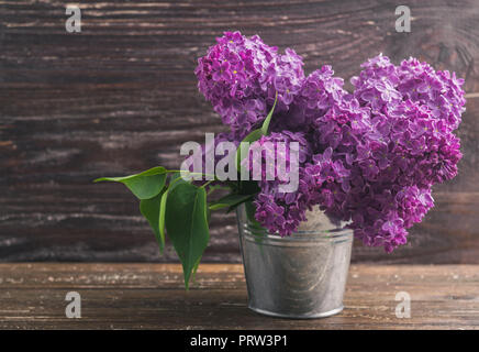 Blumenstrauß aus Lila lila Blüten in kleinen dekorativen Dose Eimer. Dunkel Braun Holz- Hintergrund. Feder romantische Blumen Dekoration. Stockfoto