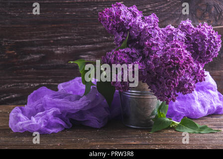 Blumenstrauß aus Lila lila Blüten in kleinen dekorativen Dose Eimer mit lila gefärbter Gaze Gewebe. Dunkel Braun Holz- Hintergrund. Feder romantische Blumen de Stockfoto