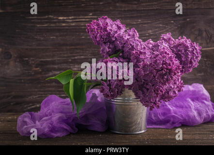 Blumenstrauß aus Lila lila Blüten in kleinen dekorativen Dose Eimer mit lila gefärbter Gaze Gewebe. Dunkel Braun Holz- Hintergrund. Feder romantische Blumen de Stockfoto