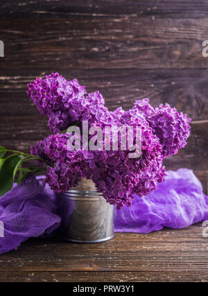 Blumenstrauß aus Lila lila Blüten in kleinen dekorativen Dose Eimer mit lila gefärbter Gaze Gewebe. Dunkel Braun Holz- Hintergrund. Feder romantische Blumen de Stockfoto