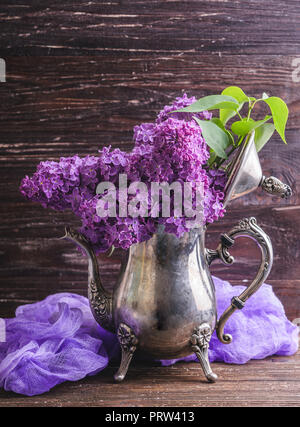 Blumenstrauß aus Lila lila Blüten in Vintage dekorative Teekanne mit lila gefärbter Gaze Gewebe. Dunkel Braun Holz- Hintergrund. Feder romantische Blumen deco Stockfoto