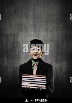 Intelligente Cute Little Boy Holding Bücher und Brille, lächelnd, während vor einem Schwarzen Brett, Stockfoto