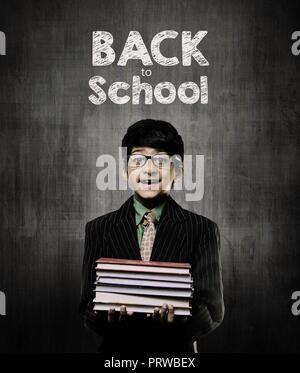 Intelligente Cute Little Boy Holding Bücher und Brille, lächelnd, während vor einem Schwarzen Brett, Zurück in die Schule an Bord geschrieben Stockfoto