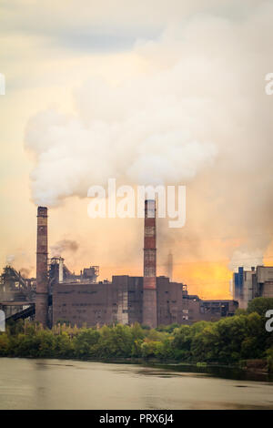 Alte Pflanze, am Ufer der Stadt platziert, mit Schornsteinen bläst Rauch in die Atmosphäre bei Sonnenaufgang Stockfoto