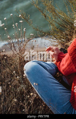 Einem jungen Rebellen sorglose Mädchen mit Zigarette im roten Pullover Rauchen in der Nähe von Meer in der goldenen Sonnenaufgang. Stockfoto