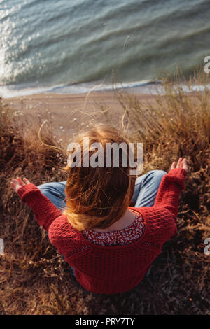 Eine junge ruhig Ginger girl in einem roten Pullover meditieren auf einer felsigen Küste mit schöner Blick aufs Meer. Stockfoto