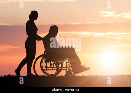 Silhouette einer Frau Unterstützung ihren behinderten Vater auf Rollstuhl im Park bei Sonnenuntergang Stockfoto