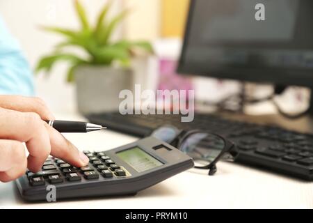 Der Mensch ist mit Taschenrechner mit Stift in der Hand. Bild von Computer, Tastatur, Blumentopf und Gläser auf dem Tisch. Stockfoto