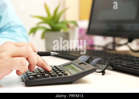 Der Mensch ist mit Taschenrechner mit Stift in der Hand. Bild von Computer, Tastatur, Blumentopf und Gläser auf dem Tisch. Stockfoto